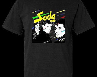 May include: Black t-shirt featuring a graphic design with the word "Soda Stereo" in yellow, with colorful lines. The design includes three black and white portraits of people.