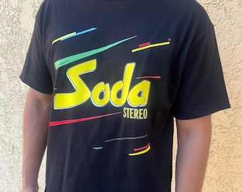 May include: Black t-shirt with the word "Soda" in large yellow letters with a white outline. The word "Stereo" is printed below the word "Soda". The shirt has colorful lines in green, blue, and red.
