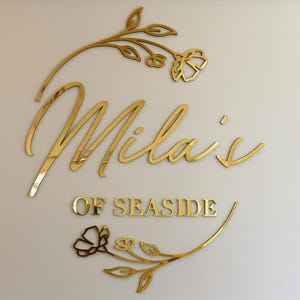 May include: Gold-colored wall art with the name "Mila's" in cursive script, and "OF SEASIDE" in block letters. Floral accents frame the text, creating a decorative and elegant design. The art is mounted on a light-colored wall.