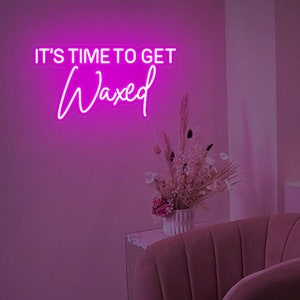 May include: Pink neon sign with the text "It's time to get waxed" in a cursive font. The sign is mounted on a pink wall with a pink couch and a vase of dried flowers in the background.