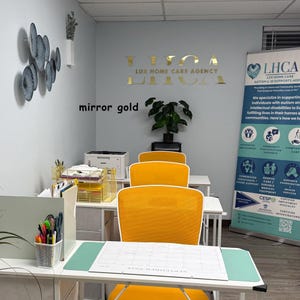 以下が含まれることがあります： 壁に「LUX HOME CARE AGENCY」と書かれた金色の文字があるオフィスインテリア。黄色い椅子、白い机、カレンダーがいくつかあります。LHCAのバナーが見えます。「mirror gold」という言葉も壁にあります。