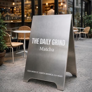 Könnte beinhalten: Ein A-förmiger Gehwegaufsteller aus gebürstetem Metall mit weißem Text, der THE DAILY GRIND, Matcha und 1234 MAIN ST, SANTA MONICA, CA 90401 lautet. Das Schild hat ein dreieckiges Profil mit bogenförmigen Ausschnitten an der Unterseite.