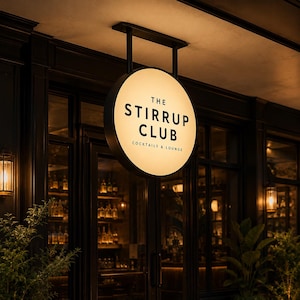 Puede incluir: Un letrero circular iluminado con marco negro que cuelga de un soporte de metal negro. El letrero presenta el texto THE STIRRUP CLUB en letras negras en negrita, con las palabras COCKTAILS & LOUNGE impresas en texto negro más pequeño.