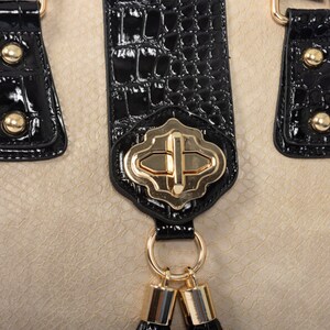 Pu&ograve; includere: Primo piano di una borsa beige con dettagli neri. La borsa presenta un motivo strutturato, manici neri con finiture dorate e una chiusura dorata. Piccoli nappe nere pendono dalla chiusura, aggiungendo un tocco decorativo al design.