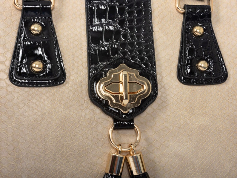 Pu&ograve; includere: Primo piano di una borsa beige con dettagli neri. La borsa presenta un motivo strutturato, manici neri con finiture dorate e una chiusura dorata. Piccoli nappe nere pendono dalla chiusura, aggiungendo un tocco decorativo al design.