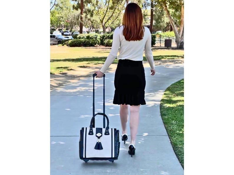 Pu&ograve; includere: Una borsa tote con ruote con un design in bianco e nero, un manico nero e un nappa nera. La borsa viene trascinata lungo un vialetto pavimentato.