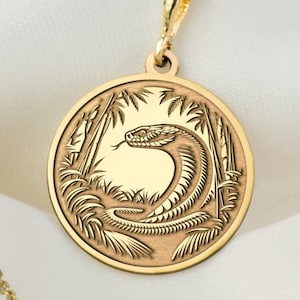 以下が含まれることがあります： 金色のペンダントネックレス。円形のフレームの中に、詳細なコブラのデザインが施されています。コブラは様式化された葉に囲まれています。ネックレスチェーンとペンダントは金色で、白い布の背景に配置されています。