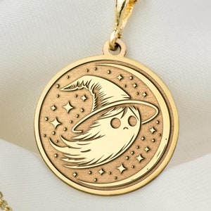 Può includere: Una collana d'oro con un ciondolo rotondo che presenta un'illustrazione stilizzata di un fantasma che indossa un cappello da strega e vola nell'aria. Il ciondolo è circondato da stelle.