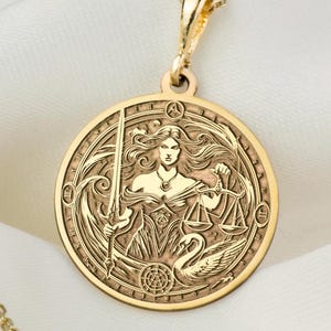 May include: Gold necklace with a round pendant featuring a detailed engraving of a woman holding a sword and scales of justice. The pendant is surrounded by a circular design.