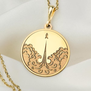 Puede incluir: Un collar de oro con un colgante redondo que presenta un cohete lanzándose a través de las nubes. El colgante está detallado con un diseño de nubes texturizado.