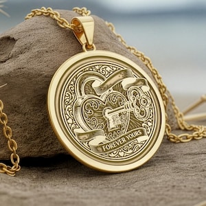 以下が含まれることがあります： 丸い彫刻が施されたチャームが付いた、ゴールドカラーのペンダントネックレス。チャームには、ハート、鍵、スクロールのデザインと「FOREVER YOURS」の文字が施されています。ネックレスチェーンもゴールドで、流木の上に置かれています。