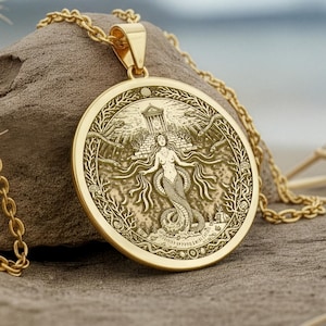 May include: A gold-colored pendant necklace featuring a detailed mermaid design. The round pendant, with a textured surface, is suspended from a delicate chain. The mermaid is depicted with flowing hair and a fishtail, surrounded by intricate details.