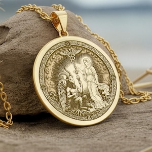 May include: A gold pendant necklace with a detailed relief design. The round pendant shows classical figures, including an angel and a woman. The gold chain is visible, and the pendant rests on a piece of driftwood. The necklace is a decorative item.