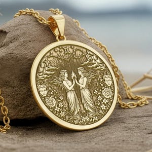 May include: A gold-colored pendant necklace featuring a detailed relief of two figures, possibly representing Gemini, surrounded by floral and celestial elements. The pendant hangs from a delicate gold chain, set against a natural, textured background.