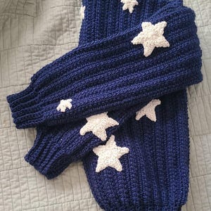 May include: A navy blue knitted sweater with white crocheted stars. The sweater has a ribbed texture and a round neckline.