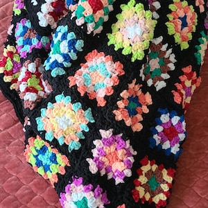 May include: A handmade, black crochet blanket featuring colorful floral squares. The squares are in shades of pink, blue, green, yellow, and orange. The blanket is a cozy, decorative item.