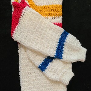 May include: White crocheted sweater with colorful stripes in red, yellow, and blue. The sweater is folded in half, showing the stripes on the sleeves.