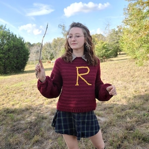 May include: A woman wearing a maroon sweater with a gold "R" on the front, a plaid skirt, and black tights. She is holding a stick in her right hand and is standing in a field.