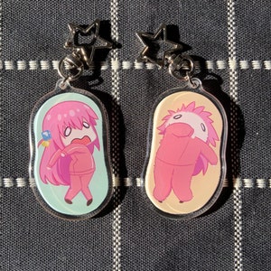 Può includere: Due portachiavi in acrilico trasparente con personaggi dei cartoni animati rosa. Un portachiavi presenta un personaggio rosa con lunghi capelli rosa e una gemma blu sulla testa. L'altro portachiavi presenta un personaggio rosa con una testa soffice rosa e bianca.