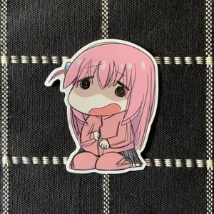 Può includere: Un adesivo con un personaggio anime dai capelli rosa con un'espressione preoccupata. Il personaggio indossa una giacca rosa e una gonna nera.