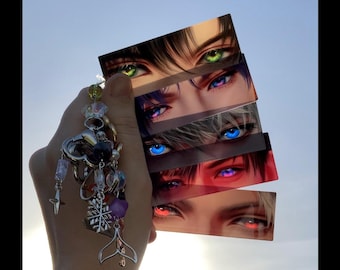 Könnte beinhalten: Eine Sammlung rechteckiger Kunstdrucke mit detaillierten Illustrationen von Augen im Anime-Stil in verschiedenen Farben, darunter Grün, Lila, Blau und Rot. Die Drucke werden mit einem dekorativen Schlüsselanhänger mit Charms zusammengehalten.