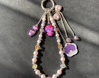 May include: A silver keychain with a beaded charm. The charm features pink pearls, clear and iridescent beads, and dangling charms with cartoon characters and pink gem accents. The keychain is on a black background.