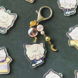Puede incluir: Un charm de acrílico transparente con un personaje de gato blanco, un cierre dorado y un colgante de cuentas. El colgante incluye cuentas rosas, naranjas y doradas, y un pequeño dije de media luna dorada. Se ven otros dijes.