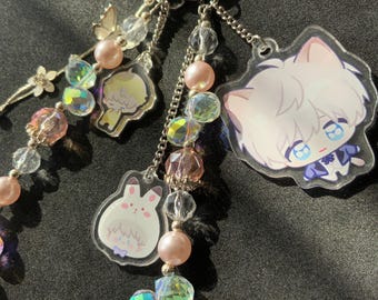 May include: A silver keychain with multiple charms. The charms include a white cat, a bunny, and a yellow figure. The keychain also features clear and iridescent beads, pink pearls, and silver chains. The background is dark.