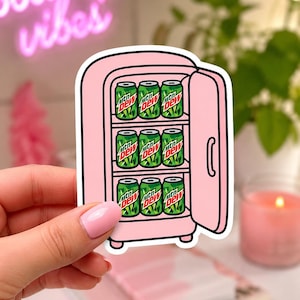 Könnte beinhalten: Ein pinkfarbener Aufkleber eines Mini-Kühlschranks mit offener Tür, der sechs Dosen Mountain Dew zeigt. Die Dosen sind grün mit dem Mountain Dew-Logo. Der Kühlschrank ist pink und hat einen Griff. Im Hintergrund befindet sich ein pinkfarbenes Neon-Schild mit der Aufschrift "good vibes".