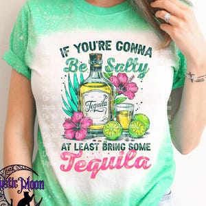 May include: A green and white t-shirt with the text "IF YOU'RE GONNA Be Salty AT LEAST BRING SOME Tequila". The graphic includes a tequila bottle, shot glass, limes, and flowers.