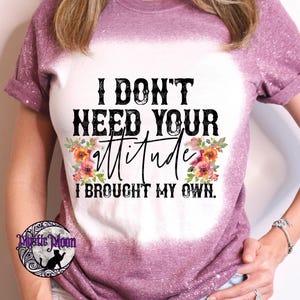 May include: A mauve short-sleeved t-shirt with a bleached white center panel. The shirt features black text that reads I DON'T NEED YOUR ATTITUDE I BROUGHT MY OWN, accented by colorful floral illustrations on the left and right sides of the word attitude.