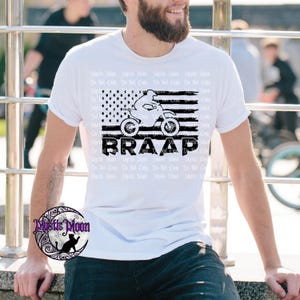 May include: White t-shirt featuring a black and white graphic of a motorcycle and rider, set against a stylized American flag. The word "BRAAP" is printed below the graphic. The shirt is a casual wear item.