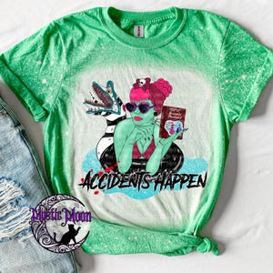 May include: A bright green bleached t-shirt featuring a graphic of a green-skinned character with pink hair, heart-shaped sunglasses, and a book titled Handbook for the Recently Deceased, next to a striped creature, with the text ACCIDENTS HAPPEN.