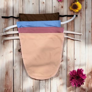 May include: Four fabric items in shades of brown, blue, mauve, and peach, each with a curved bottom edge and attached straps. The items are displayed on white hangers against a wooden background, with a few flowers.