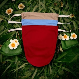 May include: Three fabric pouches in red, light blue, and tan, each with white straps. The pouches are arranged on a bed of green grass with small white and yellow flowers and two small green purses decorated with flowers.