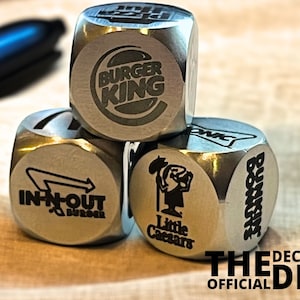 May include: Three silver metal dice with different fast food logos. The top die has the Burger King logo, the middle die has the In-N-Out Burger logo, and the bottom die has the Little Caesars logo. The dice are on a beige surface with the text "THE DECISION DICE OFFICIAL" below them.