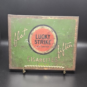 May include: Vintage Lucky Strike Cigarettes sign with a green background and a red and black circular logo. The sign reads "flat fifties" and "Cigarettes". The sign is displayed on a gold metal stand.