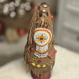 Puede incluir: Objeto decorativo en forma de cono con exterior de ante marrón, adornado con abalorios rojos, amarillos y blancos. Presenta flecos y un medallón central de cuentas con un diseño de girasol. Se ven detalles metálicos plateados.