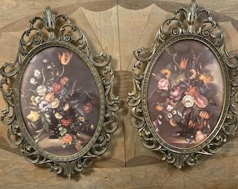 May include: Two antique oval picture frames with ornate bronze-colored metalwork. Each frame holds a floral still life painting with a dark background. The flowers are a mix of red, white, orange, and purple.
