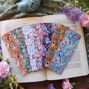 May include: A set of seven colorful bookmarks featuring a birdhouse theme. The bookmarks are printed with various floral patterns and images of birds and birdhouses. The bookmarks are laid out on an open book with a light blue bird figurine on the cover.