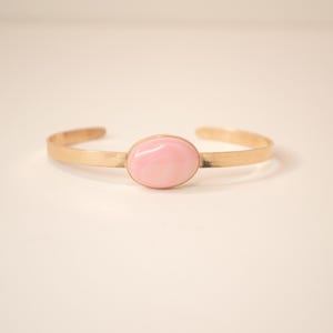 May include: A gold-colored cuff bracelet with a light pink oval stone in the center. The bracelet has a simple, elegant design, perfect for everyday wear. The stone is smooth and polished, adding a touch of color.