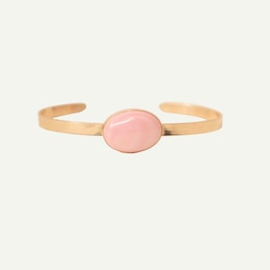 Può includere: Un bracciale rigido in metallo color oro con una pietra liscia di colore rosa chiaro di forma ovale al centro. Il bracciale è sottile e aperto, caratterizzato da una finitura metallica lucida che contrasta con la consistenza morbida e opaca.