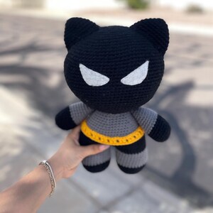 Puede incluir: Mu&ntilde;eca de gato de ganchillo negra y gris hecha a mano con detalles de ojos blancos y un cintur&oacute;n amarillo. La mu&ntilde;eca tiene un tema de superh&eacute;roe, con apariencia de gato y cuerpo a rayas. La mu&ntilde;eca es sostenida por una persona.