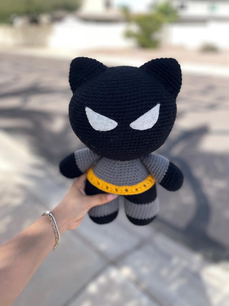 Puede incluir: Mu&ntilde;eca de gato de ganchillo negra y gris hecha a mano con detalles de ojos blancos y un cintur&oacute;n amarillo. La mu&ntilde;eca tiene un tema de superh&eacute;roe, con apariencia de gato y cuerpo a rayas. La mu&ntilde;eca es sostenida por una persona.