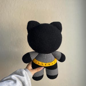 Puede incluir: Mu&ntilde;eca de gato de ganchillo negro y gris hecha a mano con cintur&oacute;n amarillo. El gato tiene una cabeza redonda con orejas puntiagudas, patas y brazos a rayas grises y negras, y un cintur&oacute;n amarillo alrededor de la cintura. La mu&ntilde;eca es suave y afelpada.