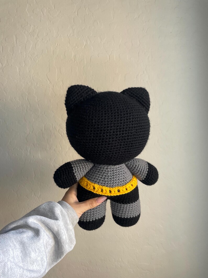 Puede incluir: Mu&ntilde;eca de gato de ganchillo negro y gris hecha a mano con cintur&oacute;n amarillo. El gato tiene una cabeza redonda con orejas puntiagudas, patas y brazos a rayas grises y negras, y un cintur&oacute;n amarillo alrededor de la cintura. La mu&ntilde;eca es suave y afelpada.