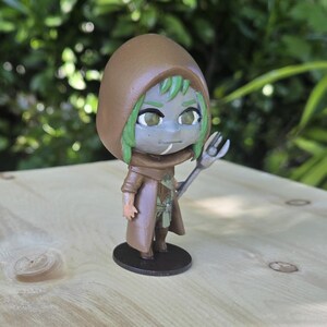 May include: A small figurine of a character with green hair, a brown hooded cloak, and a staff. The figure is standing on a wooden surface with a black base. The background features green foliage and a tree trunk.