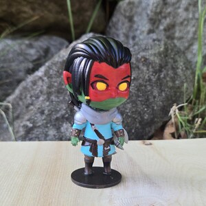 May include: A figurine of a fantasy character with red skin, green face, and yellow eyes. The figure wears a blue tunic, silver armor, and black boots. It stands on a black circular base. The background includes rocks and greenery.