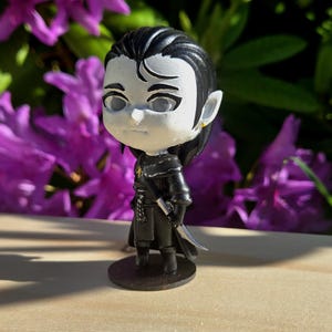 May include: A small, stylized figurine of a character with pale skin, black hair, and pointed ears. The figure is dressed in a black outfit and stands on a round base. The background features purple flowers and green foliage.