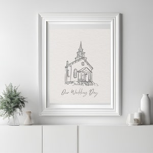 Op de afbeelding: Een zwart-witte lijntekening van een kerk met de tekst "Our Wedding Day" eronder. De tekening is ingelijst in een wit frame en hangt aan een witte muur.